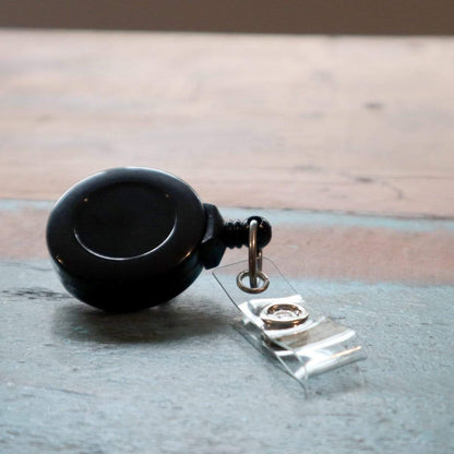 A black No Twist Badge Reel with a Swivel Spring Clip (P/N 2120-764X) lies on a wooden surface, ideal for the medical industry. Sold by Specialist ID.