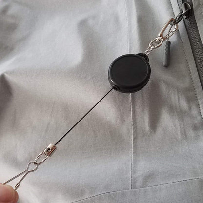 Close-up of a Ski Pass Holder Retractable Heavy Duty Sport Reel (P/N 2120-4101), clipped to a zipper on a grey fabric surface.