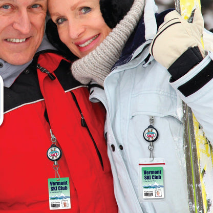 Two people, dressed warmly in winter clothing, are wearing Ski Pass Holder Retractable Heavy Duty Sport Reel (P/N 2120-4101) passes securely attached to heavy duty retractable sport reels around their necks. They are smiling, standing close together, and appear to be enjoying a snowy environment.