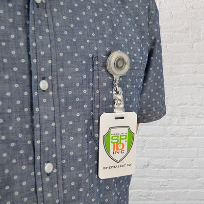 A person is wearing a blue, polka-dotted shirt with a Translucent Retractable Badge Reel with Non-Swivel Spring Clip (P/N 2120-473X) clipped to the front pocket containing an ID card that reads "SPECIALIST ID INC.