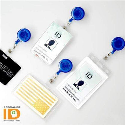 Four ID badges with Translucent Retractable Badge Reels with Non-Swivel Spring Clip (P/N 2120-473X), including two with photos, one with a gold flag design, and a black one—all labeled "Specialist ID Inc." on a white surface. The badge reel retractable cord ensures convenient access to your ID badge holder.