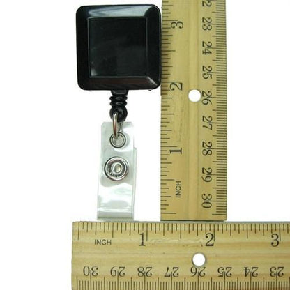 A Black Square Badge Reel With Spring Clip (P/N 2120-5701) with a retractable nylon cord is measured against a wooden ruler in inches, showcasing its clear plastic spring clip for secure attachment.