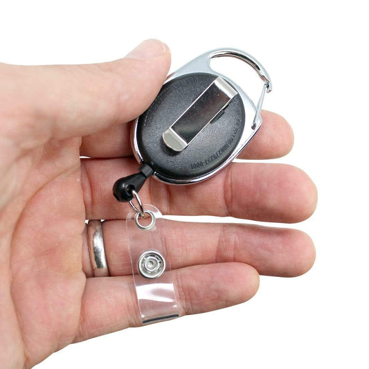 A hand holds a Premium Oval Badge Reel with Carabiner and Belt Clip (2120-71XX) with a clear plastic card holder and a metallic clip on the back, showcasing sleek sophistication. Sold by Specialist ID.