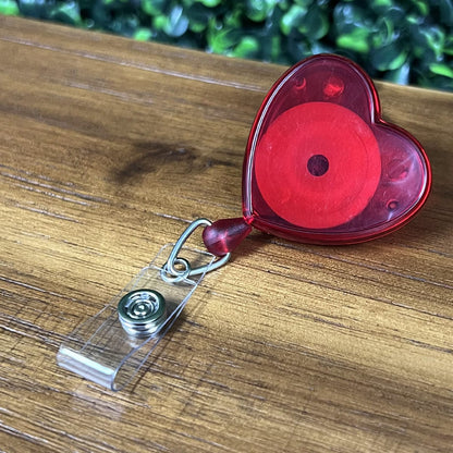 A Heart Shaped Badge Reel With Rotating Spring Clip (P/N 2120-761X), placed on a wooden surface, with green foliage in the background.