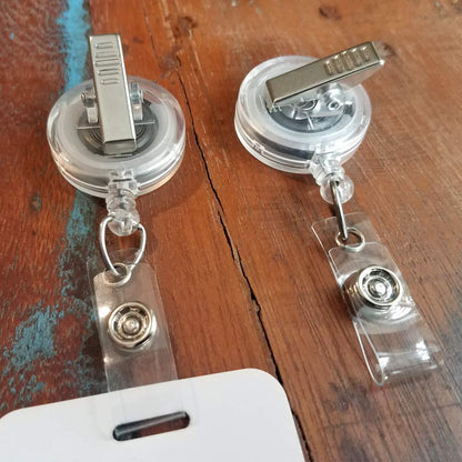 Two Translucent Badge Reels with Swivel Clip (P/N 2120-762X) with metal belt clips are placed on a wooden surface. One features a swivel clip and is positioned clip-side up, while the other is clip-side down. A badge is partially visible at the bottom.