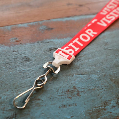 A Red Printed Visitor Lanyard With Swivel Hook (P/N 2138-5220) with the word "VISITOR" in white text, featuring a metal swivel hook and a safety breakaway clasp at the end, placed on a textured wooden surface.