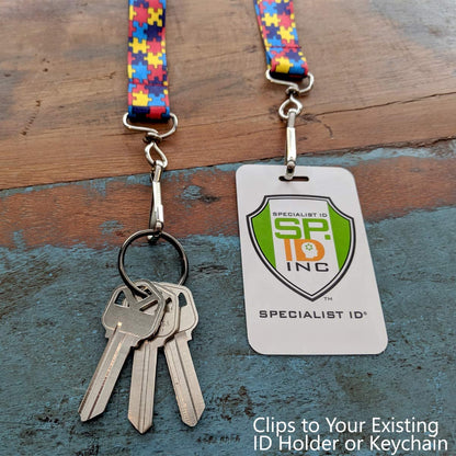 Keys attached to an Autism Awareness Flat Breakaway Lanyard With Swivel Hook (2138-5281, 2138-5282) featuring a colorful puzzle piece pattern and an ID holder with the Specialist ID logo, placed on a weathered wooden surface. Text reads: "Clips to Your Existing ID Holder or Keychain.