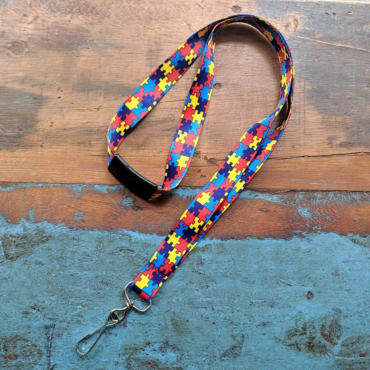 An Autism Awareness Flat Breakaway Lanyard With Swivel Hook (2138-5281, 2138-5282) with a colorful puzzle piece pattern, featuring a sturdy swivel hook at the end, is laid on a weathered wooden and blue surface.