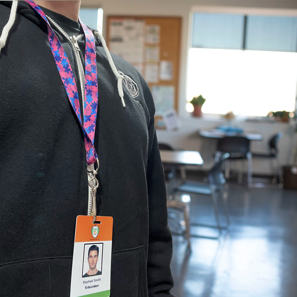 A person wearing a black hoodie with an Autism Awareness Flat Breakaway Lanyard With Swivel Hook (2138-5281, 2138-5282) displaying a badge labeled "Raphael Simh, Educator" stands in a classroom with desks and a window in the background.