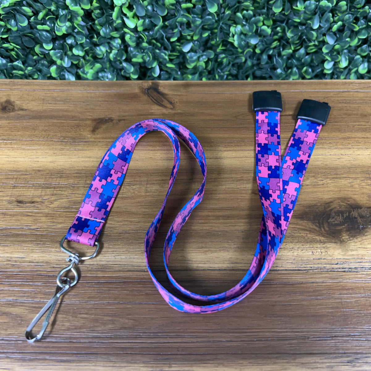 An Autism Awareness Flat Breakaway Lanyard With Swivel Hook (2138-5281, 2138-5282) with a blue and pink puzzle piece pattern is laid out on a wooden surface with a green leafy background. It features a silver swivel hook clasp.