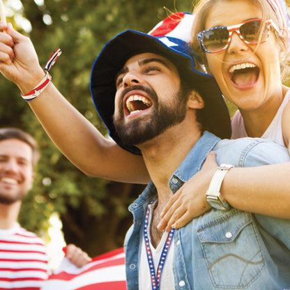 Two people smiling and cheering, one wearing a blue star-patterned hat and the other with star-shaped sunglasses, sporting 4th of July Patriotic USA Lanyard with Breakaway - Red White & Blue with Stars (2138-5305) with an American flag in the background.