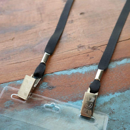 A close-up image of a black Double Clip Lanyard with 2 Bulldog Clips - Flat, Soft Material Neck Straps for Large Badge Holder Credentials (2140-530X) with gold-colored dual bulldog clips attached to a transparent plastic ID badge holder, placed on a wooden surface.