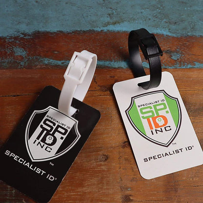 Two luggage tags, one black with a white strap and one white with a black strap, both labeled "Plastic Luggage Strap with Post and Notch -Textured (2430-201X)" and bearing a shield logo, rest on a distressed wooden surface. Each features durable travel straps for secure attachment.