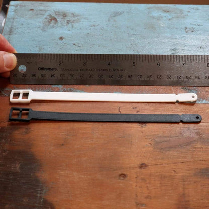 Two plastic zip ties, one white and one black, are measured with a 12-inch metal ruler on a wooden surface. A hand holds the ruler steady, highlighting their potential as **Plastic Luggage Strap with Post and Notch -Textured (2430-201X)** for securing luggage effortlessly.