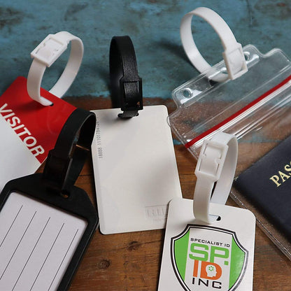 Assorted luggage tags and badges with a Plastic Luggage Strap with Post and Notch -Textured (2430-201X), including a visitor badge, a blank white card, an ID card with the text "SP INC," and a passport on a blue and wooden surface.