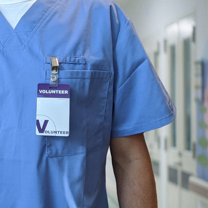 A person wearing blue scrubs with a 5 Pack - Purple Heavy Duty Plastic Volunteer Badges with Clothing Friendly Clip clipped to the front pocket stands in a hallway.