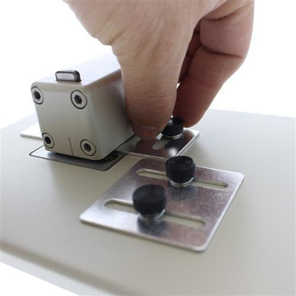 A hand is adjusting knobs on a mounted metal device. The Heavy Duty Table Top Slot Punch With Adjustable Guides (P/N SPID-9620) appears to have multiple control buttons and is secured to a flat surface with metal plates.