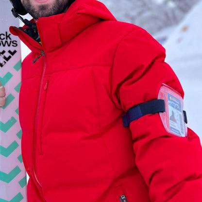 Person in a red winter jacket holding a snowboard with Vertical Armband ID Badge Holders with Zipper Top (504-ARZB & 504-ARZW) displaying a photo ID card. Snow-covered background.