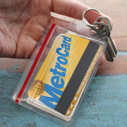 A hand holds a set of keys attached to a Clear Soft Horizontal Fuel Card or ID Badge Holder w/ Zipper Closure & Key Ring 506-ZHKR containing a yellow and blue MetroCard.