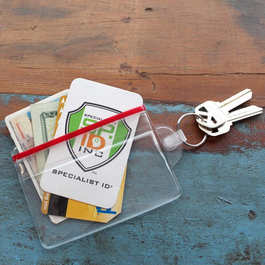 A Clear Soft Horizontal Fuel Card or ID Badge Holder w/ Zipper Closure & Key Ring 506-ZHKR containing money, cards, and a Specialist ID card is secured with a red elastic band. It rests next to keys on a blue and brown wooden surface, each key attached to a key ring.