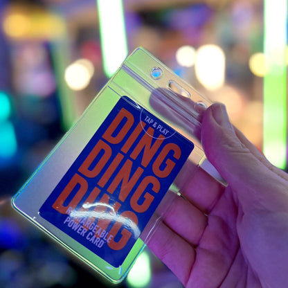 A hand holds a Clear Vertical Vinyl Badge Holder with Zipper (506-ZS-CLR) with a card inside that reads "DING DING DING RECHARGEABLE POWER CARD" in bold orange and blue text. The colorful, blurred background adds to the vibrant scene.
