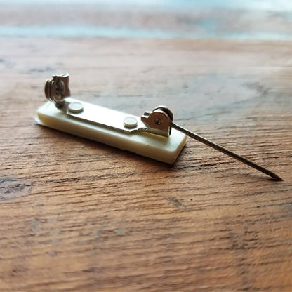 A close-up image of a Pressure-Sensitive 1 inch Nickel-Plated Steel Bar Pin with Adhesive Pin Back (P/N 5730-2620) on a wooden surface. The clasp features two fasteners and a sharp pin extended to the right, perfect for customized badges.
