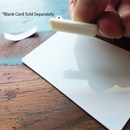 A hand holds a white clip above a blank card on a wooden surface. A small white strip lies next to the card, perfect for adding an adhesive badge pin. Text reads: "Blank Card Sold Separately. Product Name: Pressure-Sensitive 1 inch Nickel-Plated Steel Bar Pin with Adhesive Pin Back (P/N 5730-2620).