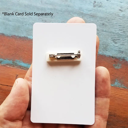 A hand holding a white card with a Pressure-Sensitive 1 inch Nickel-Plated Steel Bar Pin with Adhesive Pin Back (P/N 5730-2620) attached to it. Text on the upper left corner states "*Blank Card Sold Separately". The background consists of a surface with blue and brown tones.