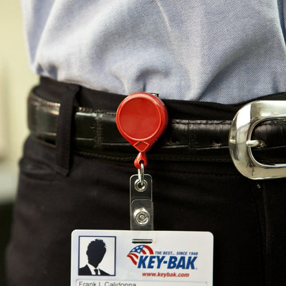A person wearing a belt with an ID badge holder clipped to it. The MINI-BAK #69 Red Retractable Reel with 36” Nylon Cord, Vinyl ID Strap and Swivel Clip 0069-005 features a red design, and a plastic badge with identification details dangles from a sturdy nylon cord.
