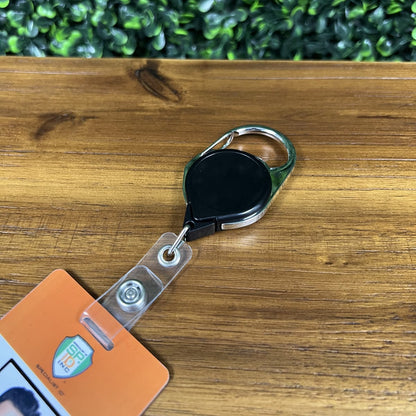 A retractable badge holder with a keyring lies on a wooden surface. An identification card is attached to the Carabiner No Twist Badge Reel (P/N 704-CB). A green leafy background is partially visible.