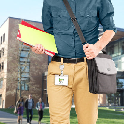 Person wearing a blue shirt and khaki pants carrying a shoulder bag with a Carabiner No Twist Badge Reel (P/N 704-CB) attached, holding folders and documents, with a campus building in the background. Other pedestrians are visible in the distance.