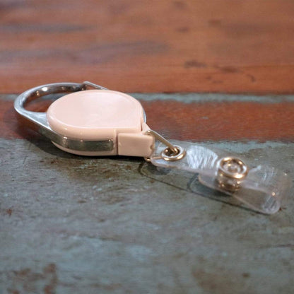A Carabiner No Twist Badge Reel (P/N 704-CB), featuring a metal clip and clear plastic strap, lies on a textured surface. Sold by Specialist ID.