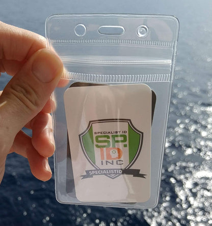 A hand holds a Clear Vertical Vinyl Badge Holder with Zipper (506-ZS-CLR) displaying a card with a logo that reads "Specialist ID, SP INC" with a ribbon at the bottom that says "SPECIALISTID". An ocean view is in the background, emphasizing the ID badge security provided by this durable vinyl badge holder.
