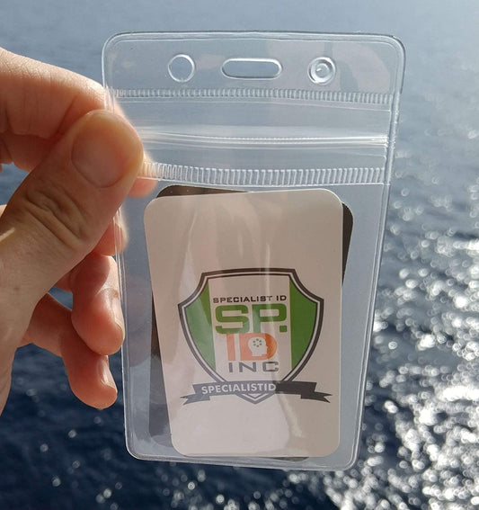 A hand holds a Clear Vertical Vinyl Badge Holder with Zipper (506-ZS-CLR) displaying a card with a logo that reads "Specialist ID, SP INC" with a ribbon at the bottom that says "SPECIALISTID". An ocean view is in the background, emphasizing the ID badge security provided by this durable vinyl badge holder.