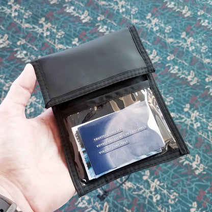 A hand holds a black Multi-Pocket Credential Neck Wallet with Lanyard (1860-300X) with a clear pocket displaying a blue card labeled "Trusted Traveler." The background features a patterned carpet with shades of blue and red.