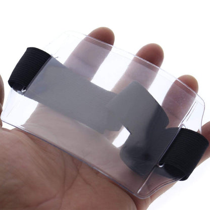 A hand holds a clear vinyl enclosure with black elastic bands at both ends, resembling Horizontal Arm ID Badge Holders (ABH-H). Sold by Specialist ID.