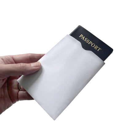 A hand holds a black passport partially inside a white paper sleeve, showcasing the SkimSAFE Shielded RFID Passport Protection Sleeve 153036 (AH-225) for identity theft prevention.