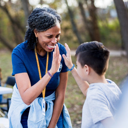 A woman in a blue shirt and a boy in a gray hoodie, both wearing 100 Pack - Anti-Bullying ½” Blue Silicone Wristband, share a high-five outdoors. Sold by Specialist ID.