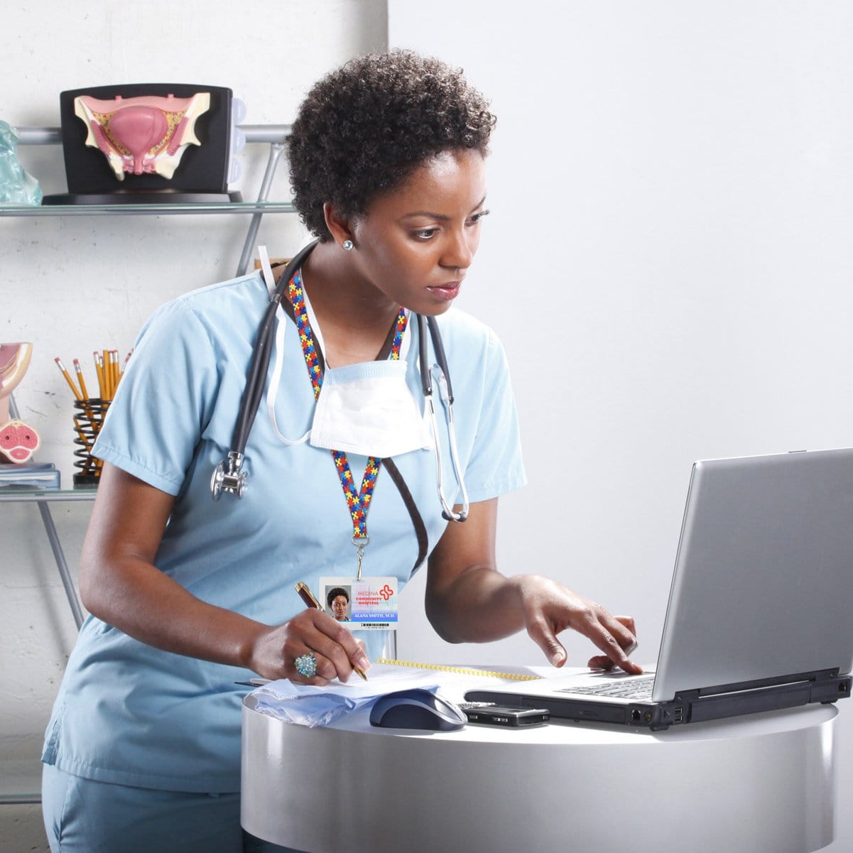 A healthcare professional in scrubs and a stethoscope, wearing an Autism Awareness Flat Breakaway Lanyard With Swivel Hook (2138-5281, 2138-5282), works on a laptop and writes on a notepad in a medical office.