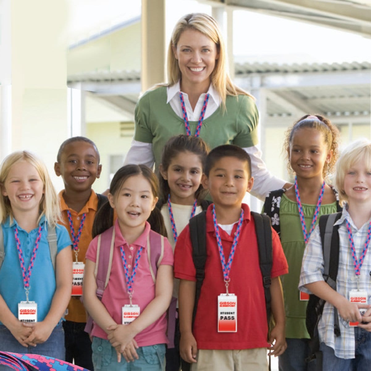A woman stands behind a group of seven smiling children, all wearing visitor passes attached to Autism Awareness Flat Breakaway Lanyard With Swivel Hook (2138-5281, 2138-5282) and backpacks, in an outdoor setting.