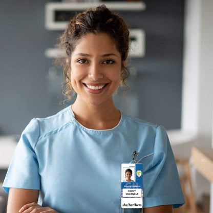 A woman in a light blue medical uniform smiles at the camera, her She/Her/Hers Vertical Pronouns Badge Buddy with White Border and laminated badge buddy displaying her commitment to diversity and inclusion.