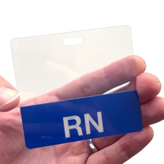 A hand holding a clear identification badge holder with a blue section at the bottom displaying the letters "RN". The Clear RN Badge Buddy - Horizontal ID Badge Backer for Nurses - Double Sided Print adds an extra layer of professionalism, making it easy to identify registered nurses.