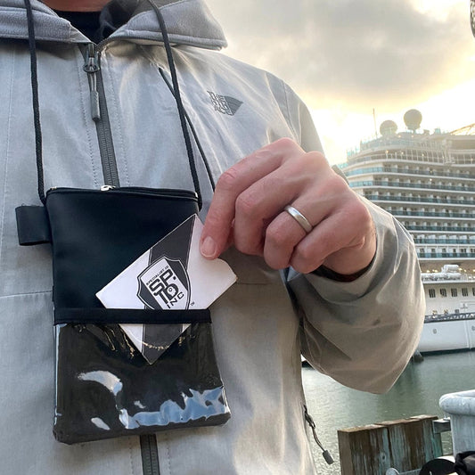A person wearing a gray jacket is holding a card with a logo and placing it into a Vinyl Credential Neck Wallet with Adjustable Lanyard and Top Zipper Pouch (CW-6-BLK) around their neck. A large ship is visible in the background.