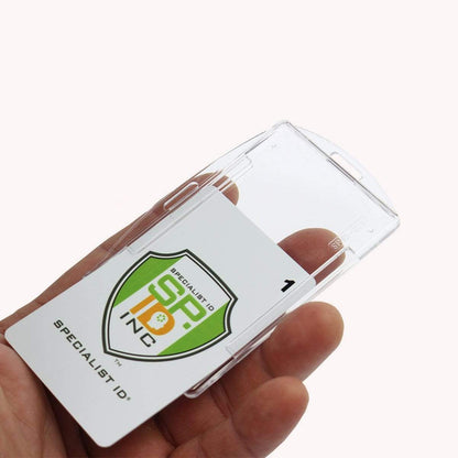 A hand holds a Crystal Clear Rigid Open-Faced Single ID Card Holder (P/N SPID-075) made from durable polycarbonate material. Inside, a card with "SP INC" and a shield logo is visible, labeled "Specialist ID" at the bottom. The adaptable slot holes allow for convenient attachment to various lanyards or clips.