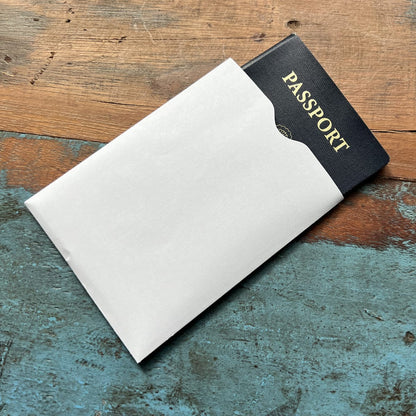 A black passport, partially inserted into a white SkimSAFE Shielded RFID Passport Protection Sleeve 153036 (AH-225), rests on a wood and blue-painted surface.