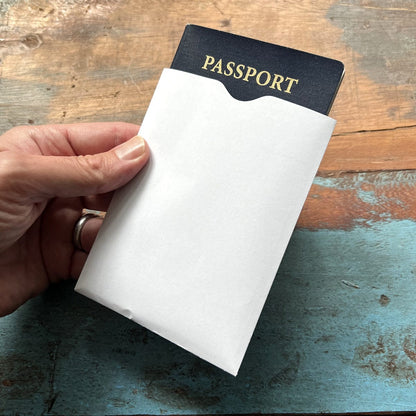 A hand holds a white envelope containing a dark-colored passport, equipped with the SkimSAFE Shielded RFID Passport Protection Sleeve 153036 (AH-225) for added identity theft prevention, on a wooden surface.