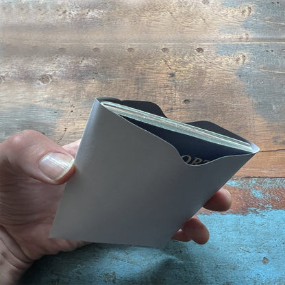 A hand holding a white envelope containing a booklet, with SkimSAFE Shielded RFID Passport Protection Sleeve 153036 (AH-225) for identity theft prevention, all set against a wooden surface in the background.