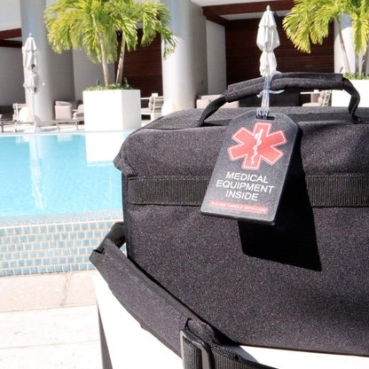 A black medical equipment bag with a red "2 Pack - Heavy Duty Medical Equipment Luggage Tags CPAP / BIPAP with Plastic Loops" is placed near a pool surrounded by white lounge chairs and palm trees.