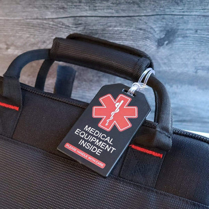 A black bag with a 2 Pack - Heavy Duty Medical Equipment Luggage Tags CPAP / BIPAP with Plastic Loops displaying a medical symbol and text that reads "Medical Equipment Inside - Please Handle With Care" is shown against a grey background.