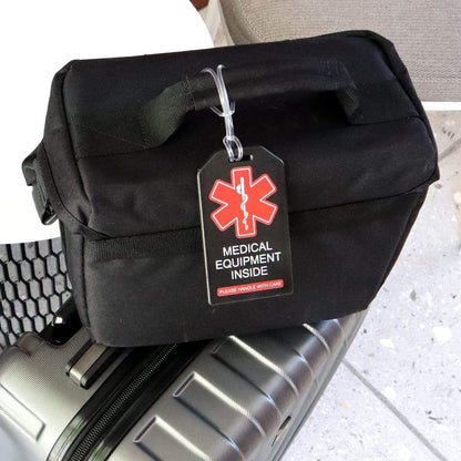 A black bag with a 2 Pack - Heavy Duty Medical Equipment Luggage Tags CPAP / BIPAP with Plastic Loops reading "Medical Equipment Inside, Please Handle With Care" is placed atop a silver suitcase.